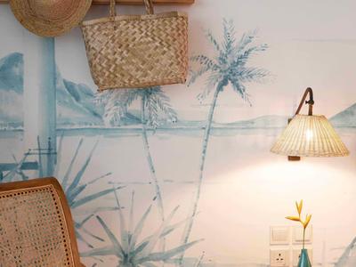 Heller Raum mit Bambusstuhl, Holzregal, Strohhut, Tasche und Wandlampe über Holztisch mit Vase.