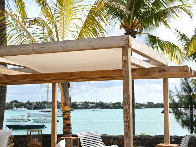 Freiluftterrasse mit Stühlen und Tischen unter einem Holzpavillon mit Palmen und Meer im Hintergrund.
