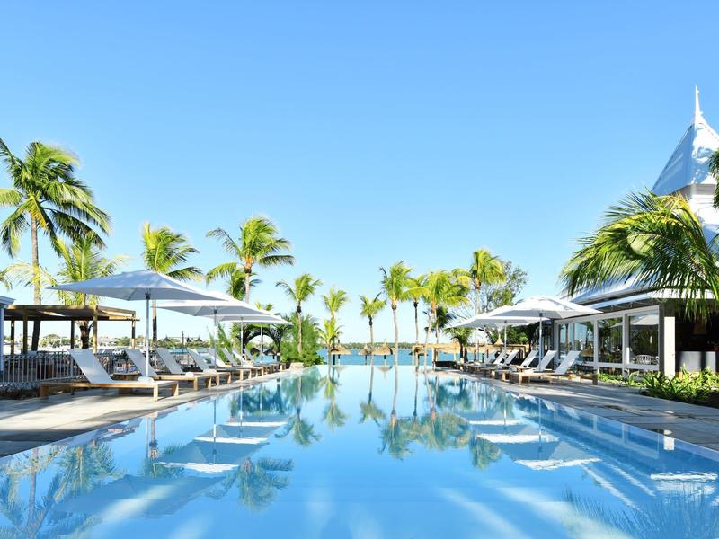 Infinity-Pool mit weißen Sonnenschirmen und Liegestühlen umgeben von Palmen unter klarem blauem Himmel