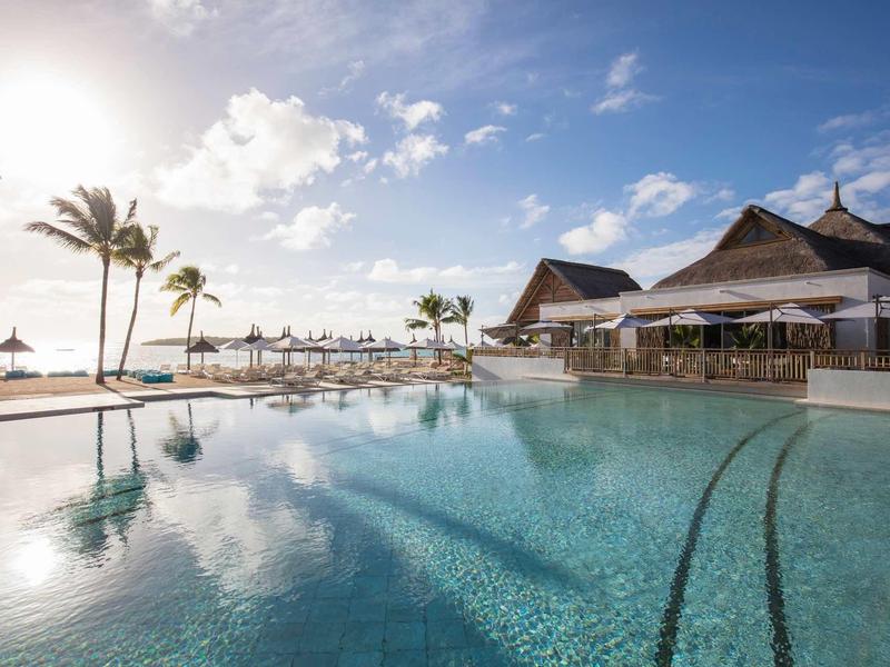 Großer Pool mit klar klarem Wasser, Palmen und Sonnenschirmen, blauem Himmel und Sonne im Hintergrund.
