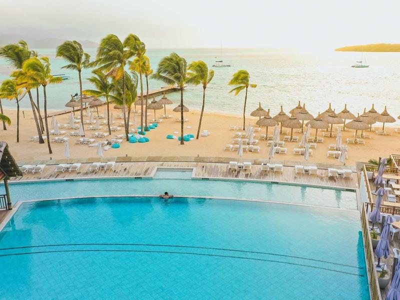 Großer Pool mit Sonnenliegen, Palmen und Strand mit Sonnenschirmen am Meer im Sonnenlicht.