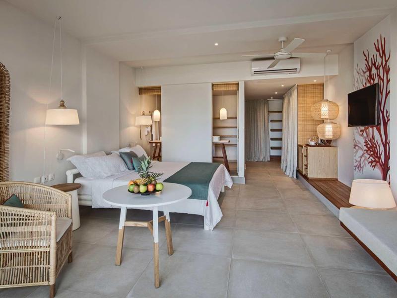 Helles Zimmer mit Doppelbett, Korbstühlen, Fernseher an Wand und Zugang zu begehbarem Kleiderschrank.