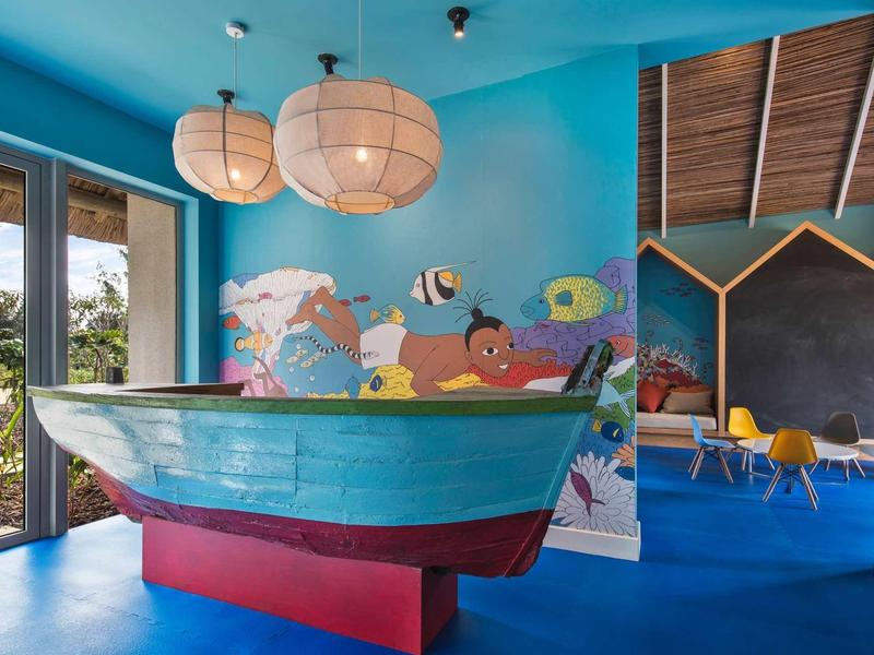 Ein Kinderzimmer mit blauer Wand, Boot-Bett, bunter Fische-Wanddeko und großen Fensterflächen.