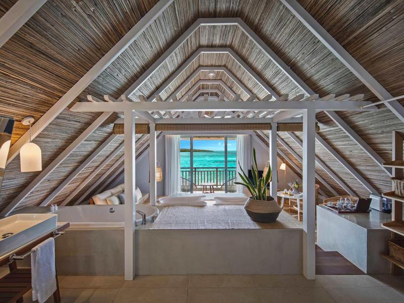 Offenes Dachzimmer mit Holzbalken, großem Bett, Pflanzen und Meerblick durch Fenster.