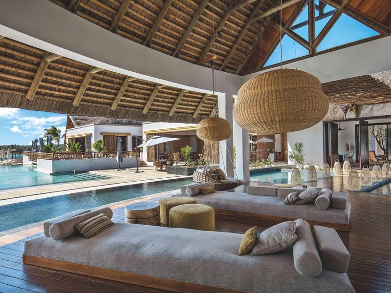 Offener Lounge-Bereich mit großen Hängelampen, Holzdach und Pool im Hintergrund bei sonnigem Wetter.