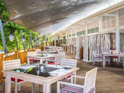 Ristorante luminoso e aperto con sedie e tavoli in legno su una terrazza accanto a piante verdi.