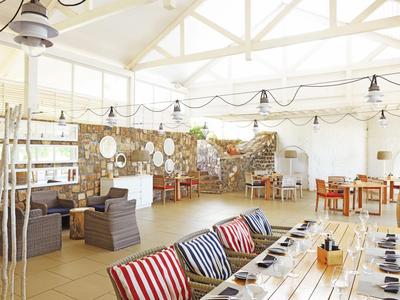 Caffè luminoso con mobili in legno, cuscini colorati e pareti in pietra rustiche sotto un soffitto con travi bianche.