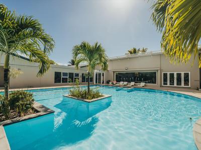 Zona de piscina con palmeras y tumbonas frente a un edificio de hotel bajo un cielo azul.