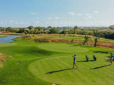 Tres personas juegan al golf en un campo bien cuidado con un estanque y árboles al fondo.