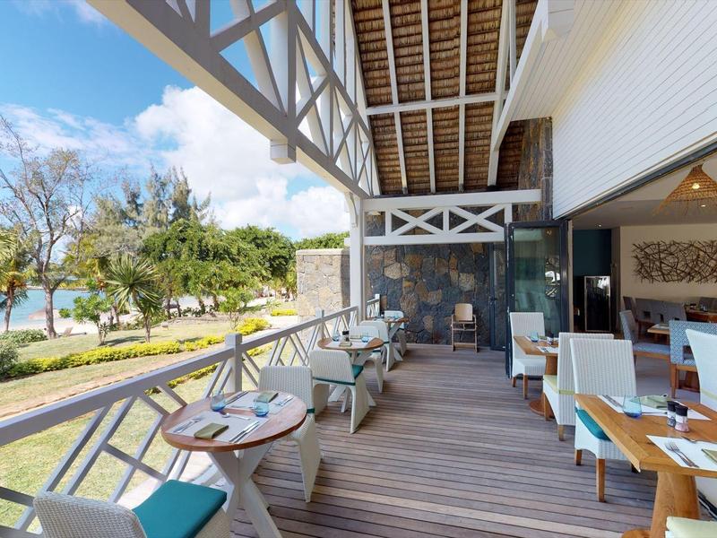 Terraza abierta con áreas de asiento y mesas de comedor en un hotel con vista a plantas tropicales y cielo.