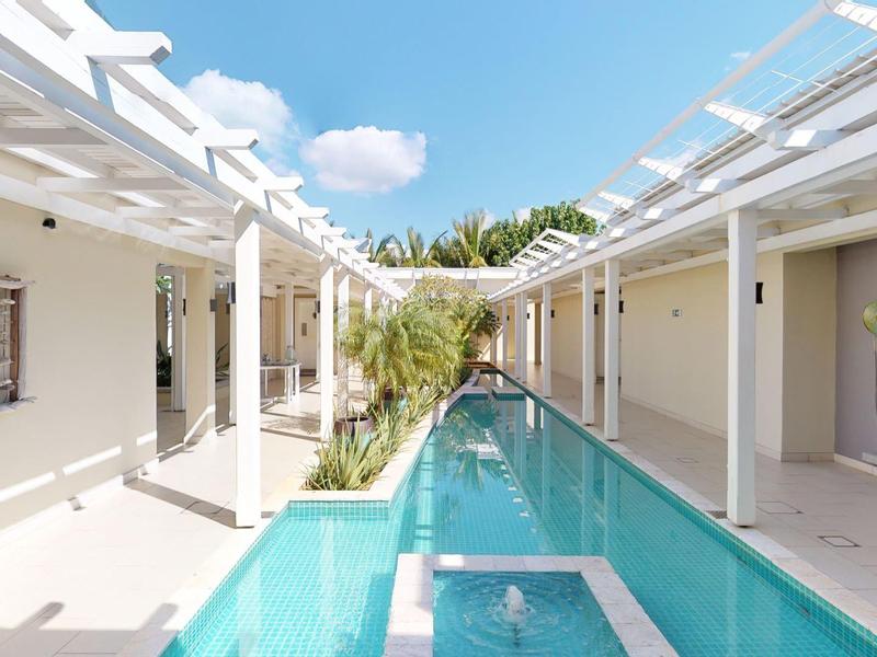 Piscina moderna de hotel con pérgola y paredes blancas, cielo soleado y vegetación al fondo.