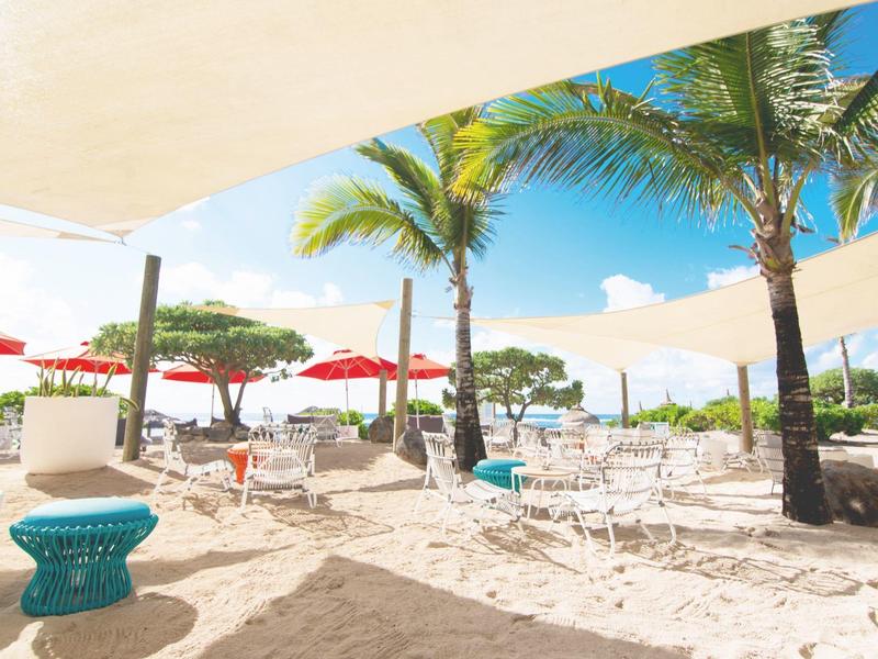 Beach resort with white sand, palm trees, umbrellas, and chairs under sunshades.