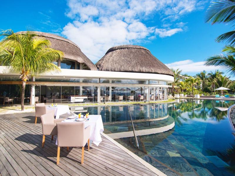Modern hotel area with tables by the pool and round thatched buildings under a blue sky.