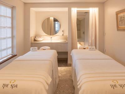 Zwei weiße Massageliegen in einem hellen, ruhigen Spa-Raum mit rundem Spiegel und beruhigender Deko.