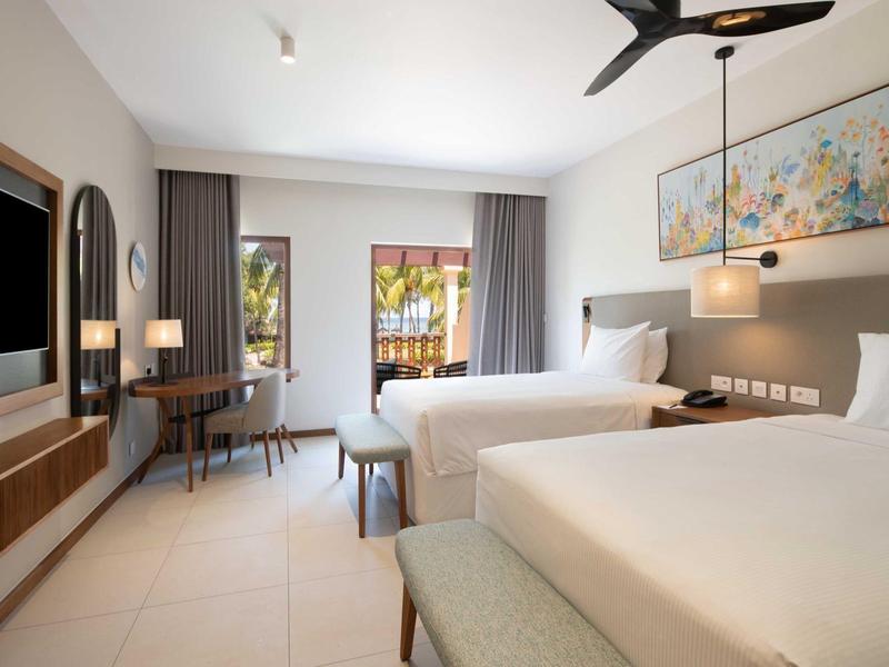 Helle Hotellzimmer mit zwei großen Betten, Schreibtisch, Fernseher und moderner Beleuchtung.