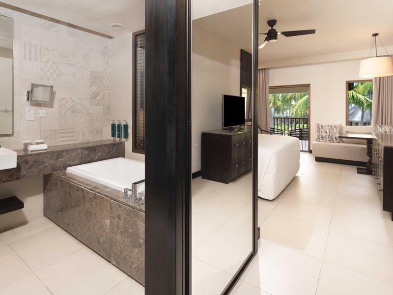 Helles Hotelzimmer mit Dusche, Badewanne, Doppelbett, Fernseher und Blick auf Balkon mit Natur.