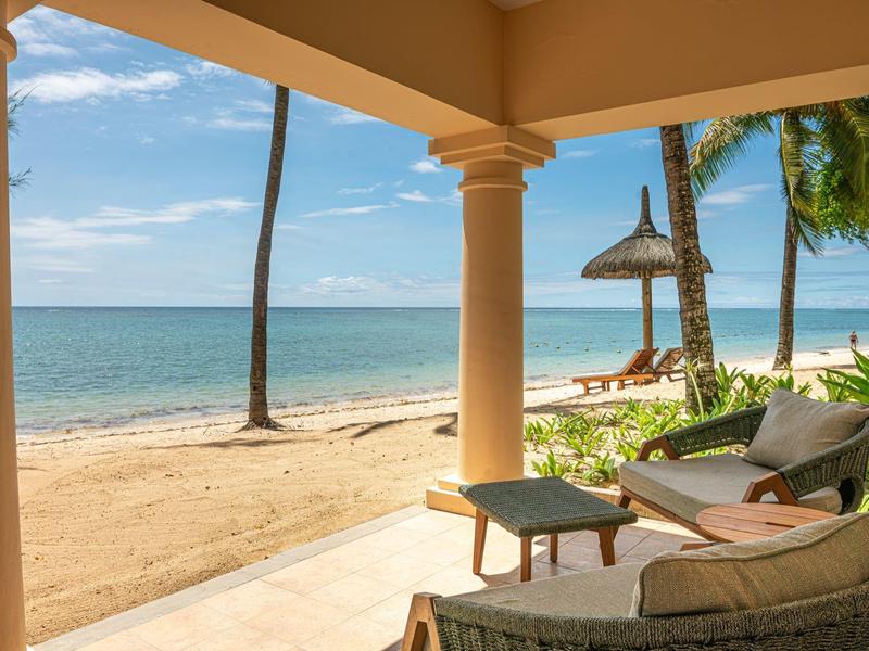 Blick von einer Terrasse mit Stühlen und Tisch auf einen Sandstrand mit Palmen und ruhigem Meer.