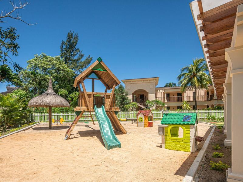 Kinder-Spielplatz mit Rutsche, Klettergerüst, Sandboden und kleinen bunten Spielhäusern unter blauem Himmel.