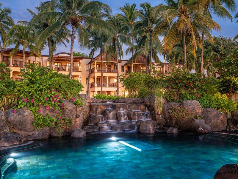 Ein tropischer Pool mit Wasserfall, umgeben von Palmen und einem hellen Resort-Gebäude im Hintergrund.