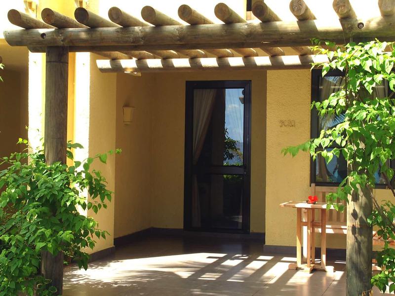 Haus mit gelben Wänden, Holzpergola und Eingang, umgeben von grünen Pflanzen und einem Tisch mit Stuhl.