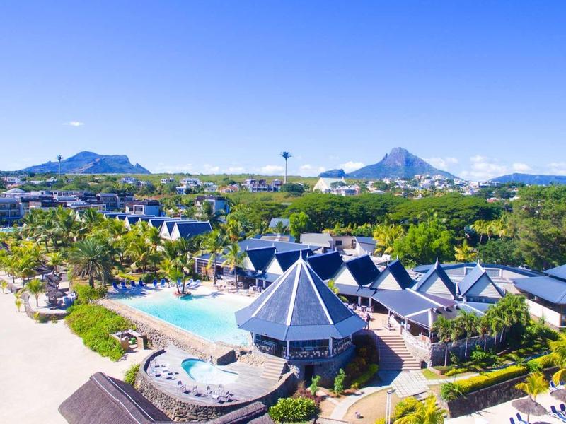 Resort mit Pool, grünen Gärten, Palmen und Bergen im Hintergrund bei klarem Himmel.