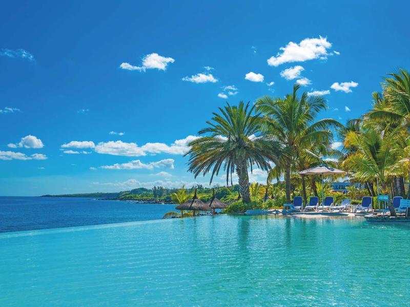 Infinity-Pool mit Palmen, blauem Himmel und Meer im Hintergrund an tropischem Ort.
