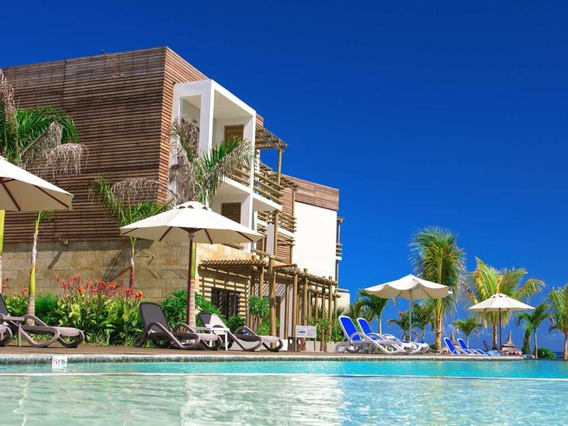 Moderner Holzbau mit Sonnenschirmen und Liegen neben klarem Pool unter blauem Himmel.