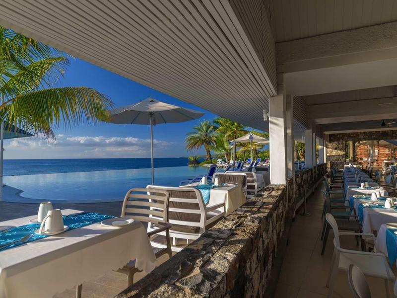 Restaurant mit Tischen, Stühlen und Poolblick bei sonnigem Himmel und Palmen am Meer.