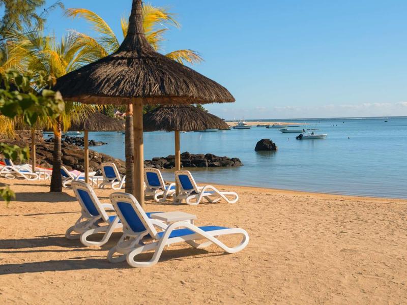 Sandstrand mit blauen Liegestühlen unter Strohdächern und Palmen, ruhiges blaues Meer im Hintergrund.