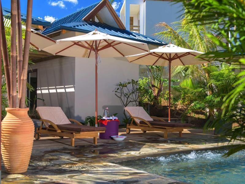 Poolbereich mit Liegestühlen unter Sonnenschirmen, umgeben von Pflanzen und blauem Himmel.
