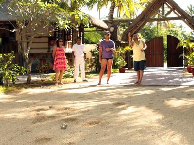 Menschen spielen Boule auf einem sandigen Platz vor einem tropischen Hotel mit Palmen.