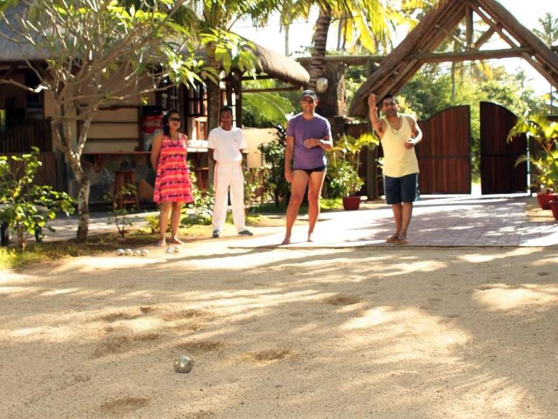 Quatre personnes jouent à la pétanque sur un sol sablonneux sous des palmiers devant un bâtiment en bois.