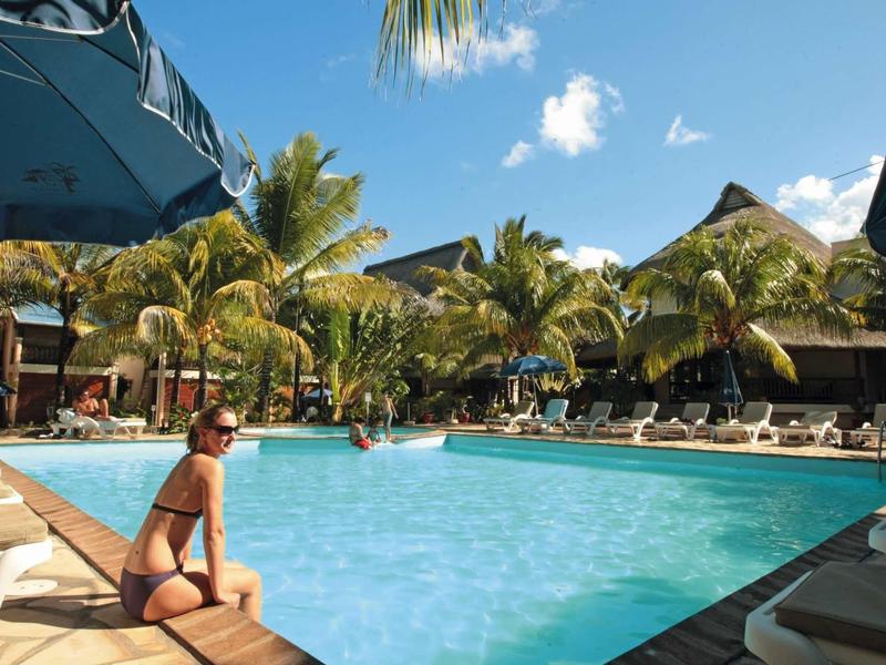 Femme se détendant au bord de la piscine d'un hôtel tropical avec palmiers et parasols.