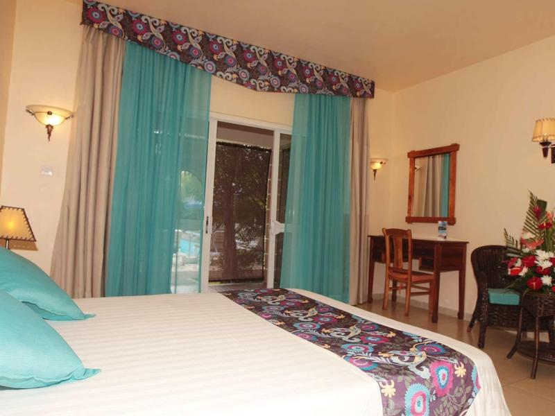 Chambre d'hôtel confortable avec grand lit, bureau et accès au balcon avec rideaux turquoise.