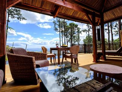 Offener Holzpavillon mit Korbsesseln und Tisch, umgeben von Bäumen und blauem Himmel.
