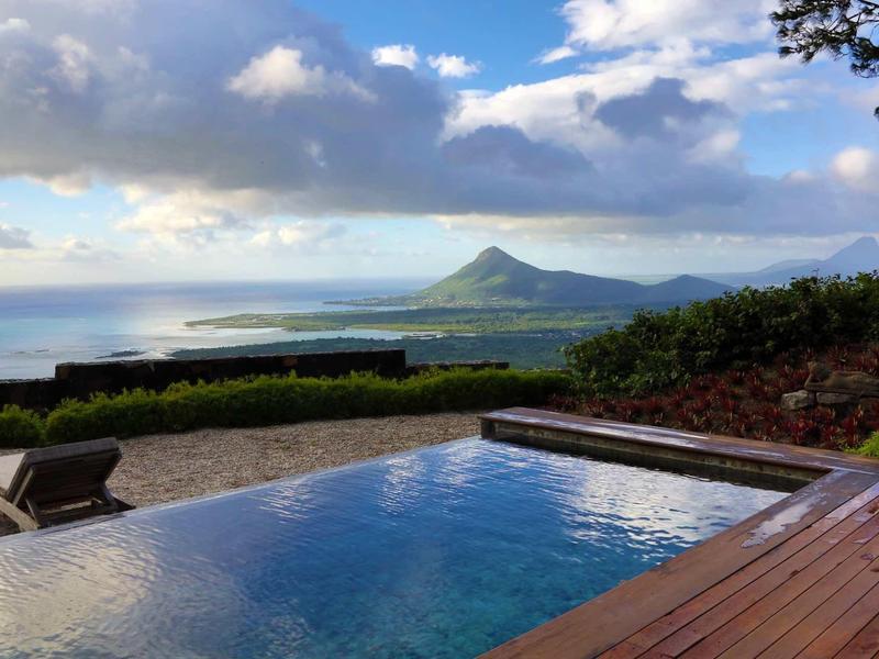Infinity-Pool mit Holzdeck, grünen Büschen, Meer und Berg unter bewölktem Himmel im Hintergrund.
