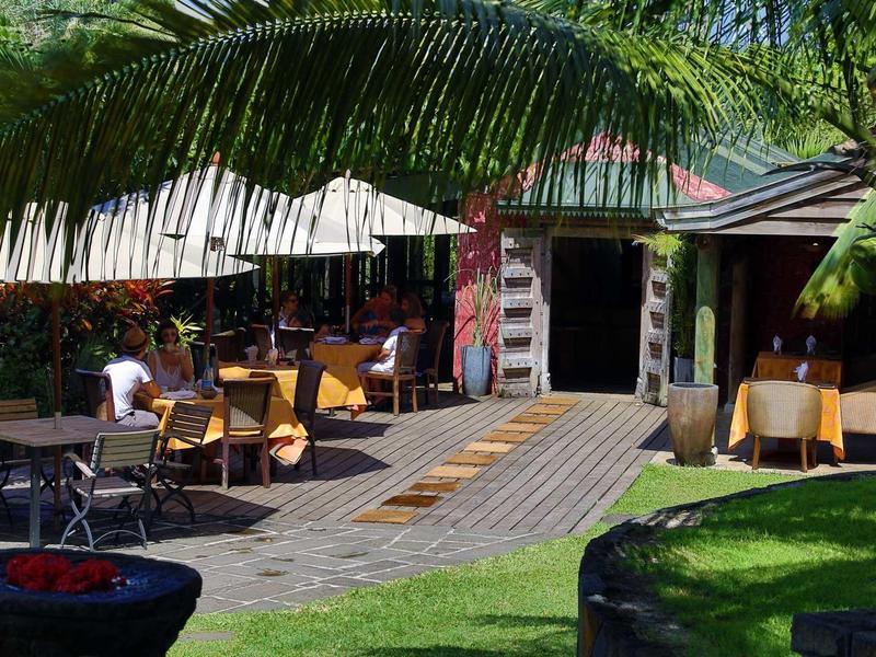 Außenbereich eines Restaurants mit Tischen, Stühlen, Sonnenschirmen und grüner Pflanzenumgebung.