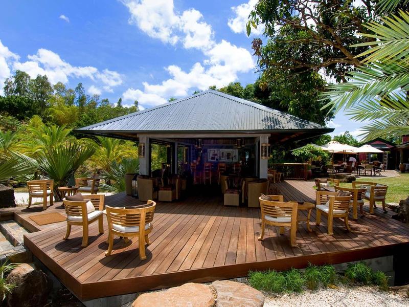 Ein Holzdeck mit runden Tischen und Stühlen vor einem kleinen Café, umgeben von Palmen und Blauem Himmel.