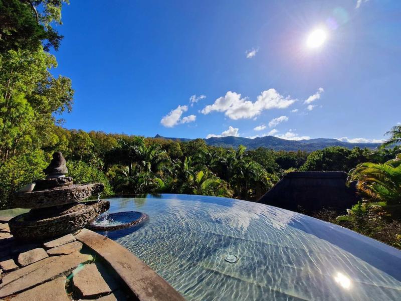 Infinity-Pool mit klarem Wasser, umgeben von grünen Bäumen und Bergen unter strahlend blauem Himmel.