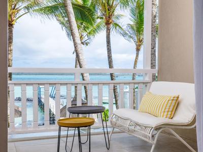 Balkon z białą sofą, żółtą poduszką i małymi stolikami z widokiem na palmy i morze.