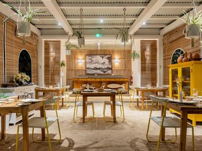 Elegantes Restaurant mit Holzmöbeln, großen Fenstern, grünen Pflanzen und warmem Licht.
