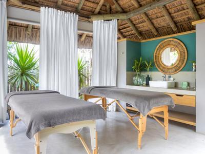 Innenraum eines hellen Spa mit zwei grauen Massageliegen, weißen Vorhängen und tropischer Dekoration.