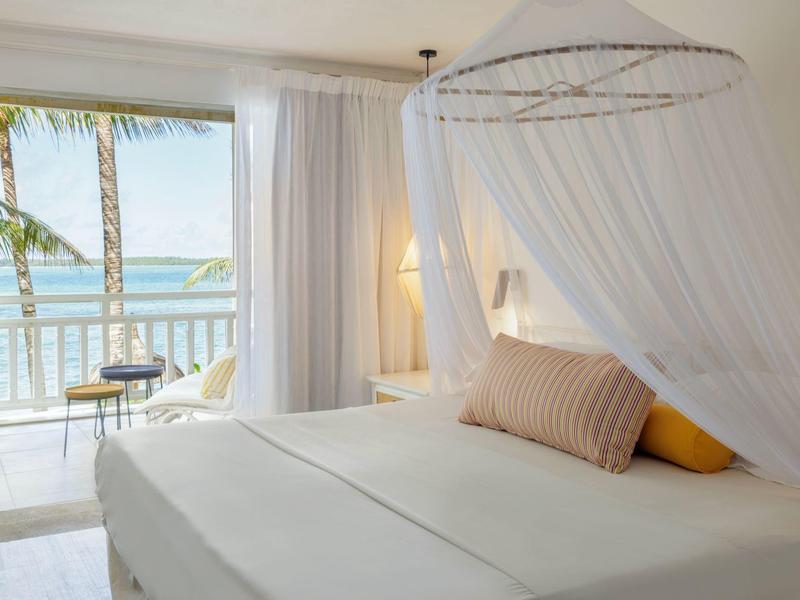 Jasny pokój z widokiem na morze, białą moskitierą i balkonem z palmami.