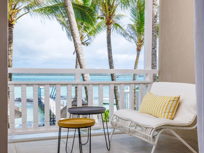 Balkon z białą sofą, żółtą poduszką i małymi stolikami z widokiem na palmy i morze.