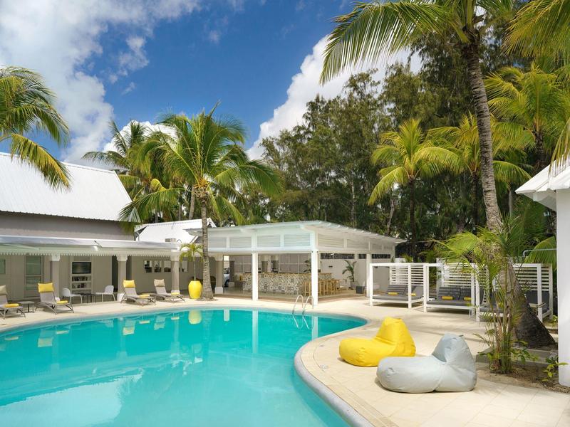 Strefa basenowa ośrodka z białymi budynkami, palmami i poduszkami przy krawędzi basenu.