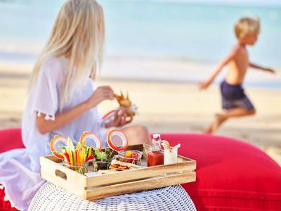 Vrouw houdt dienblad met ontbijtopties op het strand, kind rent op de achtergrond.