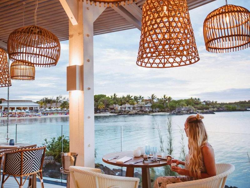 Vrouw zit in open restaurant aan het water met houten lampen en uitzicht op de baai.