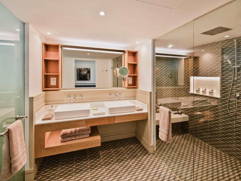 Moderne hotelbadkamer met dubbele wastafels, grote spiegelwand en inloopglazen douche.