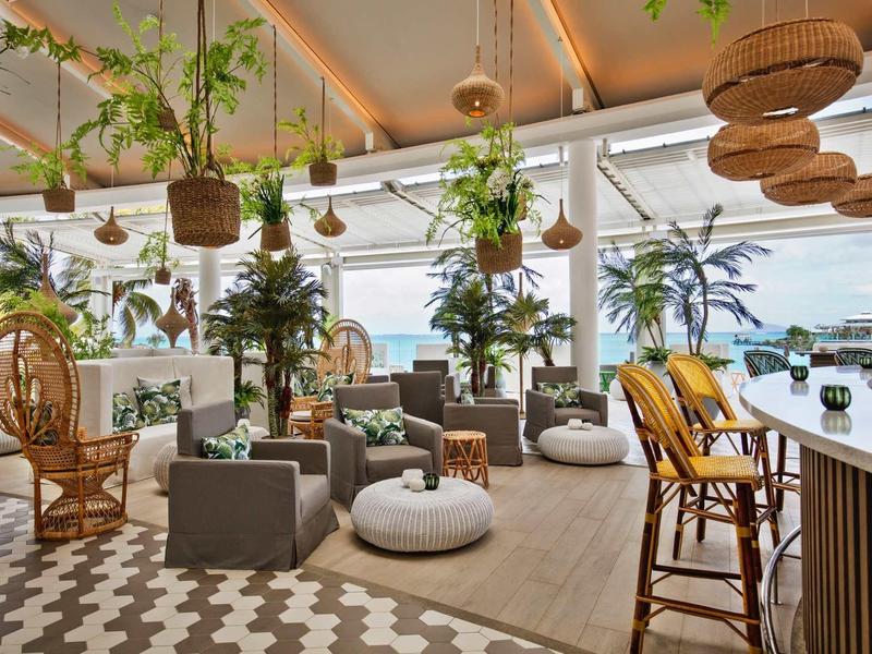 Lichte lounge met groene planten, rotan meubels en uitzicht op een haven met boten.