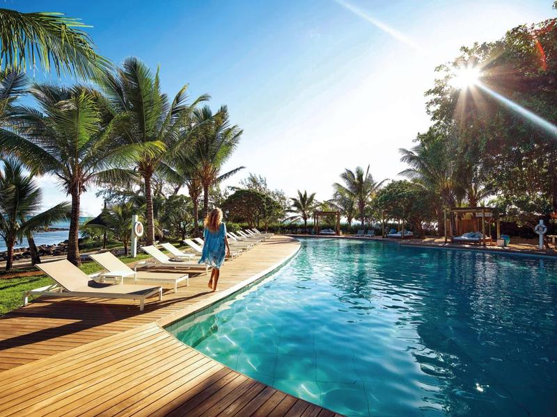 Vrouw loopt langs de rand van een tropisch zwembad met palmbomen en ligstoelen in het zonlicht.